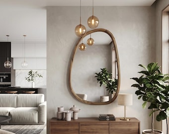 May include: An organic-shaped wall mirror with a natural wood frame hangs above a rectangular wooden console table. The mirror reflects a room with a green plant, while amber-colored glass pendant lights hang above the mirror.