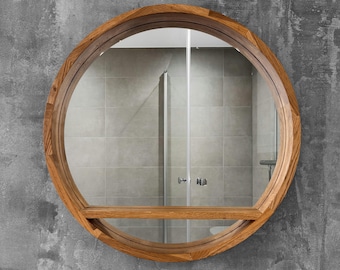 May include: A circular wall mirror featuring a thick, natural-toned wooden frame with a visible grain pattern. The mirror includes a horizontal wooden shelf integrated across the lower third of the circular glass surface, providing a flat ledge.