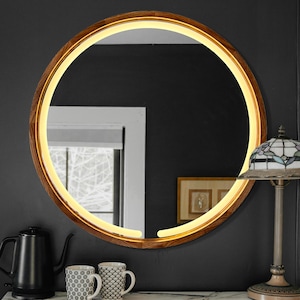 Puede incluir: Espejo redondo con marco de madera e iluminación perimetral. El marco de madera presenta un veteado natural. El espejo está fijado a una pared gris oscuro. La luz cálida crea un brillo suave.