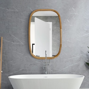 以下が含まれることがあります： グレーの壁の前に、波打つデザインの白いバスタブがあります。バスタブの上には、木製のフレームの楕円形の鏡が、クロムの蛇口と一緒に吊り下げられています。