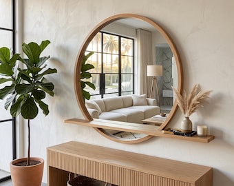 May include: A large circular wall mirror with a light wood frame and an integrated horizontal shelf extending across the lower section. The mirror reflects a living room interior, while the shelf holds a small vase with dried pampas grass.