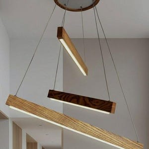 Op de afbeelding: Een plafondlamp met drie lineaire houten balken van verschillende lengtes, opgehangen aan dunne metalen kabels aan een ronde houten plafondkap. Elke balk bevat een geïntegreerde led-lichtstrip langs de onderrand.