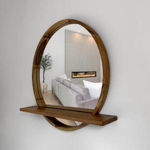 May include: A round wooden mirror with a built-in shelf. The mirror has a natural wood finish and a minimalist design. The shelf is rectangular and positioned below the mirror. The reflection shows a living room with a sofa and fireplace.