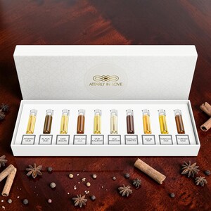 May include: A white rectangular gift box containing ten small glass vials of perfume, each labeled with names including Afghan Oud, Black Oud, and Turkish Oud. The box lid features the gold text Attarly In Love. The vials contain various shades of oil.