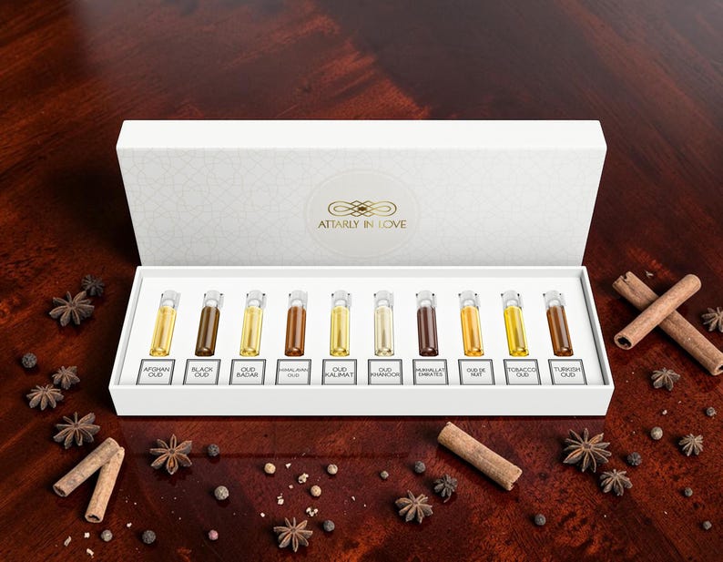 May include: A white rectangular gift box containing ten small glass vials of perfume, each labeled with names including Afghan Oud, Black Oud, and Turkish Oud. The box lid features the gold text Attarly In Love. The vials contain various shades of oil.