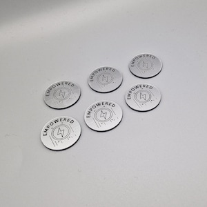 Puede incluir: Seis fichas redondas de color plateado con la palabra "EMPOWERED" grabada en el borde. Cada ficha presenta un símbolo de rayo en el centro. Las fichas tienen un borde negro y están dispuestas sobre una superficie gris claro.