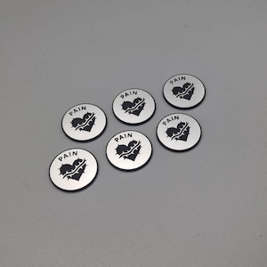 May include: Six silver-coloured round tokens are displayed. Each token has a black heart pierced with thorns and the word "PAIN" in black. The tokens are arranged on a light grey surface.