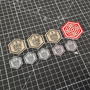 Può includere: Una collezione di gettoni di gioco esagonali con illustrazioni a tema vichingo. I gettoni sono in vari colori, tra cui beige, grigio e rosso. Alcuni gettoni hanno il numero "5" stampato sopra, mentre altri hanno il numero "1".