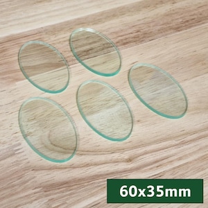 Op de afbeelding: Vijf heldere, ovaalvormige glazen stukken met een lichte groene tint liggen op een houten oppervlak. De afmetingen zijn 60x35mm. De glazen stukken zijn transparant.