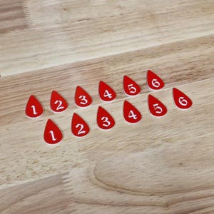 May include: Red teardrop-shaped game pieces with white numbers 1 to 6. The pieces are arranged in two rows on a wooden surface. The numbers are clearly visible against the red background.