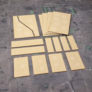 Puede incluir: Un conjunto de piezas de terreno de madera sin terminar, cortadas con láser, para juegos de mesa. El conjunto incluye varias baldosas rectangulares y cuadradas con un patrón cuadrado repetido, y una baldosa grande con una grieta. Las piezas están dispuestas sobre una superficie gris.
