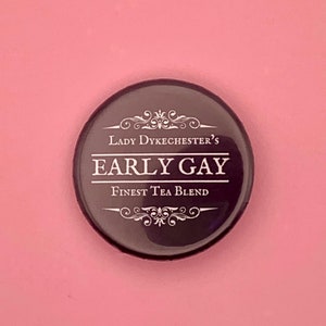 Könnte beinhalten: Ein runder, dunkelbrauner Knopf mit dem weißen Text "Lady Dykechester's EARLY GAY Finest Tea Blend". Der Knopf befindet sich auf einem rosa Hintergrund.