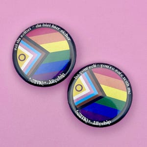 Könnte beinhalten: Zwei runde schwarze Buttons mit einem Regenbogenflaggen-Design und dem Text "be yourself - you're safe with me" und "LGBTIO*-Allyship". Die Buttons haben ein Farbschema aus Rosa, Lila, Blau, Grün, Gelb, Orange und Rot.