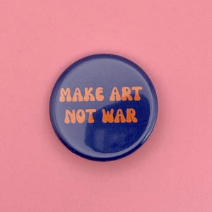 Könnte beinhalten: Ein runder, marineblauer Button mit dem orangefarbenen Text "MAKE ART NOT WAR" auf einem rosa Hintergrund. Der Button ist eine einfache, kühne Aussage.