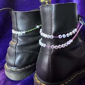 Op de afbeelding: Twee zwarte leren laarzen met kralen enkelbandjes. Eén enkelbandje spelt "ADORE" in witte kralen op een groene draad. De andere zegt "PRIDE CUTIE" in witte kralen op een roze draad, met hartvormige kralen.
