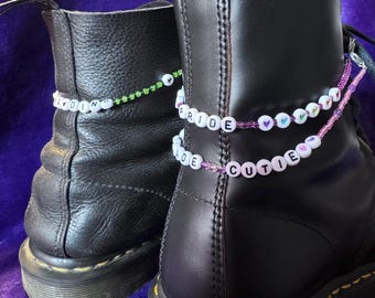 May include: Two black leather boots adorned with beaded anklets. One anklet spells out "ADORE" in white beads on a green string. The other anklet says "PRIDE CUTIE" in white beads on a pink string, with heart-shaped beads.