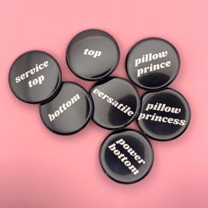 Könnte beinhalten: Sieben schwarze runde Knöpfe mit weißem Text auf rosa Hintergrund. Die Knöpfe tragen Aufschriften wie "top", "bottom", "versatile", "pillow prince", "pillow princess", "power bottom" und "service top".