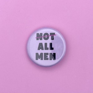 Könnte beinhalten: Ein runder, hellvioletter Button mit dem Text "NOT ALL MEN" in Schwarz, mit einem rosa Strich durch das Wort "NOT". Der Button befindet sich vor einem einfarbigen rosa Hintergrund.