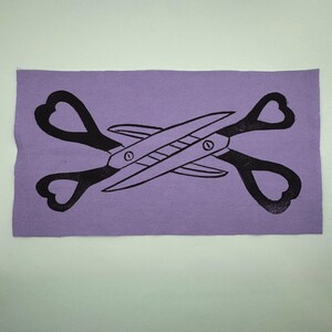 May include: A rectangular lavender fabric patch featuring a black scissors design. The scissors are crossed, with heart-shaped handles. The blades and pivot points are clearly defined, creating a bold, graphic image.