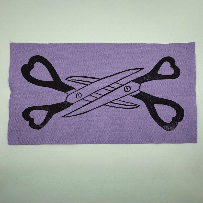May include: A rectangular lavender fabric patch featuring a black scissors design. The scissors are crossed, with heart-shaped handles. The blades and pivot points are clearly defined, creating a bold, graphic image.