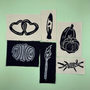 Op de afbeelding: Set van zeven zwarte en witte stoffen patches met verschillende ontwerpen, waaronder een hart, een pompoen, een lucifer, een werveling, prikkeldraad en een lava lamp.