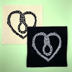 Op de afbeelding: Twee stoffen patches met een zwart hartvormig motief met een strop erin. De ene patch is wit met zwarte stiksels en de andere is zwart met zilveren stiksels.