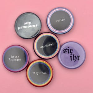 Könnte beinhalten: Eine Sammlung runder Ansteckbuttons mit Pronomen in verschiedenen Farben und Designs. Die Buttons zeigen Texte wie "any pronouns", "er/ihm", "they/them", "dey/deren", "keine Pronomen" und "sie ihr". Der Hintergrund ist einfarbig rosa.
