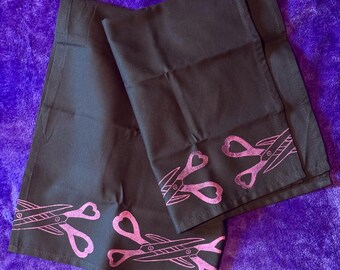 May include: Two black rectangular fabric towels featuring a repeating screen-printed design of pink scissors with heart-shaped finger loops. The scissors are positioned horizontally along the bottom edge of each towel, showcasing a clean hem.