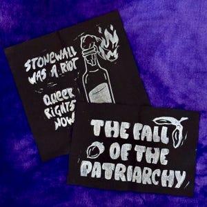 Könnte beinhalten: Zwei schwarze Stoffaufnäher mit weißem Text und Illustrationen. Ein Aufnäher lautet "Stonewall Was a Riot Queer Rights Now" mit einer Flaschenillustration. Der andere lautet "The Fall of the Patriarchy".