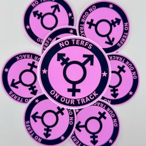 May include: A collection of pink circular stickers with a navy blue border. Each sticker features a transgender symbol in the center, surrounded by the text "NO TERFS ON OUR TRACK" and small star accents.