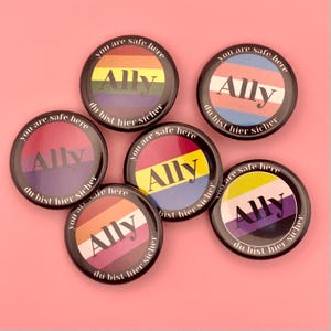 Könnte beinhalten: Eine Sammlung runder Ally-Anstecknadeln mit verschiedenen LGBTQ+-Pride-Flaggen-Designs. Jede Anstecknadel zeigt das Wort "Ally" in fetter Schrift, umgeben von den Sätzen "You are safe here" und "du bist hier sicher."