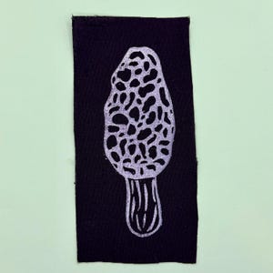 Op de afbeelding: Een zwart stoffen patch met een witte illustratie van een morielje. De paddenstoel heeft een getextureerde, putjeskap en een geribbelde steel. De patch is rechthoekig en geplaatst tegen een lichtgroene achtergrond.