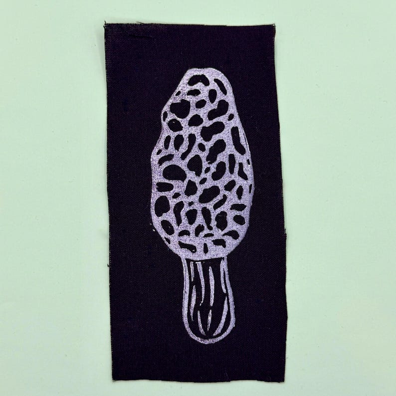 Op de afbeelding: Een zwart stoffen patch met een witte illustratie van een morielje. De paddenstoel heeft een getextureerde, putjeskap en een geribbelde steel. De patch is rechthoekig en geplaatst tegen een lichtgroene achtergrond.