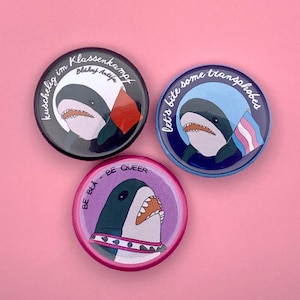 May include: Three circular badges featuring shark illustrations. One is black with German text and a shark wearing a hat with a flag. Another is blue with the text "Let's bite some transphobes" and a shark with a trans flag. The third is purple with the text "Be Bla - Be Queer".