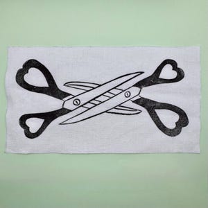May include: Black and white linocut print of scissors with heart-shaped handles, crossed over each other. The print is on a rectangular piece of white fabric, set against a light green background.