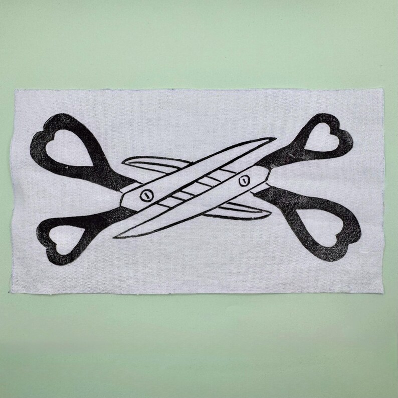 May include: Black and white linocut print of scissors with heart-shaped handles, crossed over each other. The print is on a rectangular piece of white fabric, set against a light green background.