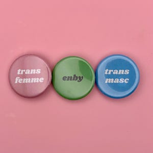Könnte beinhalten: Drei runde Buttons in Rosa, Grün und Blau. Der rosa Button trägt die Aufschrift "trans femme", der grüne Button "enby" und der blaue Button "trans masc". Die Buttons befinden sich auf einem rosa Hintergrund.