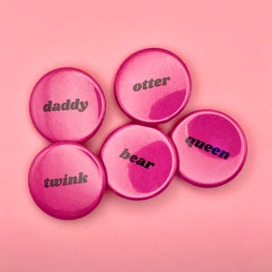 Könnte beinhalten: Fünf rosa runde Buttons mit schwarzem Text. Der Text auf den Buttons lautet "daddy", "otter", "twink", "bear" und "queen". Die Buttons sind auf einem rosa Hintergrund angeordnet.
