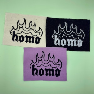 Op de afbeelding: Drie stoffen patches met het woord "homo" gedrukt in zwarte gotische letters op een witte, zwarte en paarse achtergrond. Elke patch heeft een gestileerd vlammenontwerp boven het woord.