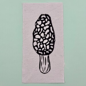 Op de afbeelding: Een zwart-wit linoleumsnede van een morielje op een rechthoekig stuk stof van natuurlijke kleur. De paddenstoel heeft een getextureerde, putjeskap en een geribbelde steel. De stof staat tegen een lichtgroene achtergrond.