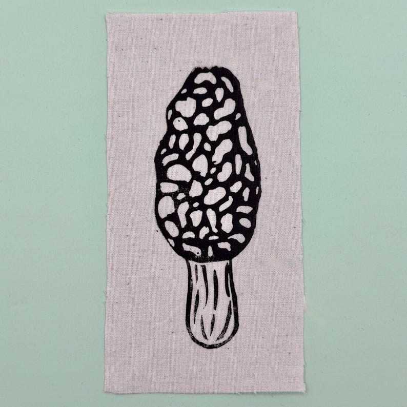 Op de afbeelding: Een zwart-wit linoleumsnede van een morielje op een rechthoekig stuk stof van natuurlijke kleur. De paddenstoel heeft een getextureerde, putjeskap en een geribbelde steel. De stof staat tegen een lichtgroene achtergrond.