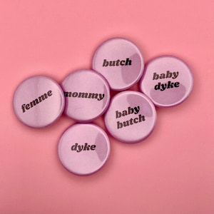 Könnte beinhalten: Sechs hellviolette runde Buttons mit schwarzem Text. Die Buttons tragen die Wörter: "butch", "baby dyke", "baby butch", "dyke", "mommy" und "femme". Der Hintergrund ist einfarbig hellrosa.