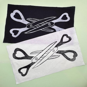 May include: Two fabric squares, one black and one white, each with a scissors design. The scissors have heart-shaped handles and a detailed blade. The design is in contrasting colours on each square.