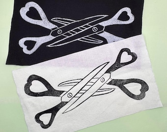 May include: Two fabric squares, one black and one white, each featuring a scissors design. The scissors have heart-shaped handles and a detailed blade. The design is in contrasting colors on each square.
