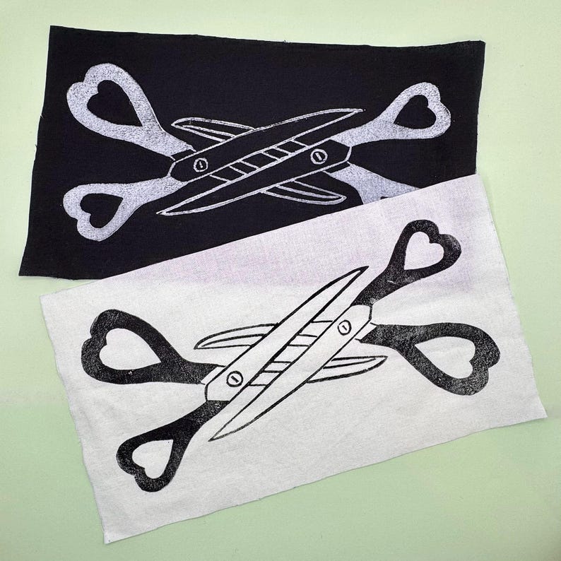 May include: Two fabric squares, one black and one white, each featuring a scissors design. The scissors have heart-shaped handles and a detailed blade. The design is in contrasting colors on each square.