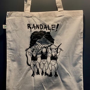 Könnte beinhalten: Eine weiße Tragetasche mit schwarzen Henkeln und einem schwarzen grafischen Aufdruck von drei Ratten auf Rollschuhen, die eine Flagge mit der Aufschrift "RANDALE!" halten.