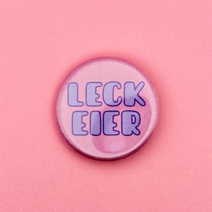 Könnte beinhalten: Ein runder Ansteckbutton mit einem verlaufenden rosa Hintergrund. Der Text LECK EIER ist in einer fetten, abgerundeten, dunkelvioletten Schrift in der Mitte des Buttons aufgedruckt. Das Design ist schlicht und der Text nimmt den Platz ein.