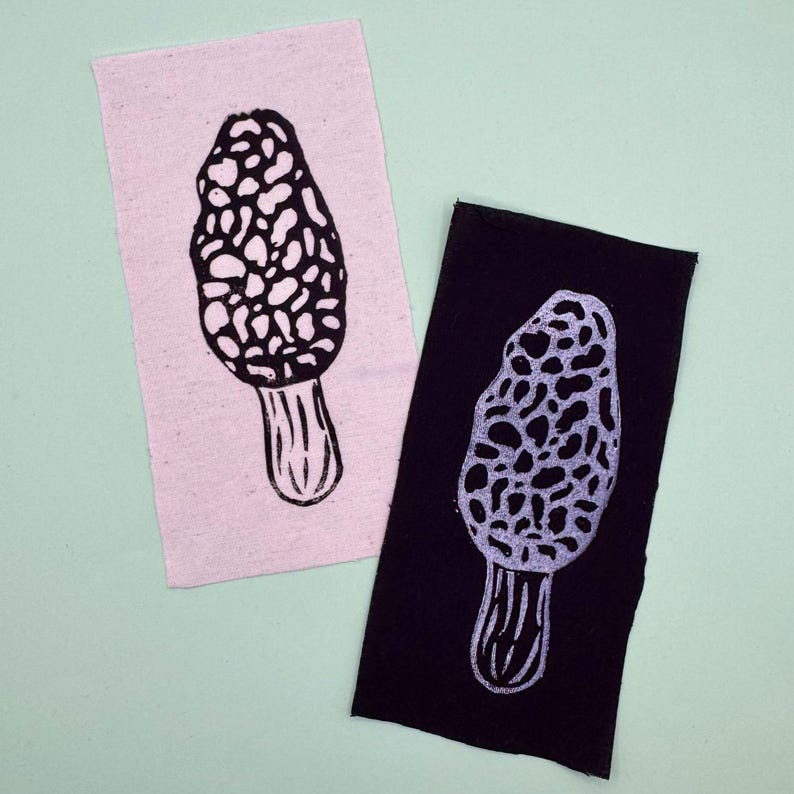 Op de afbeelding: Twee stoffen patches, &eacute;&eacute;n lichtroze en &eacute;&eacute;n zwarte, elk met een zwart-wit morel paddenstoel print. Het paddenstoelontwerp is een gedetailleerde illustratie van de getextureerde hoed en steel van de paddenstoel.