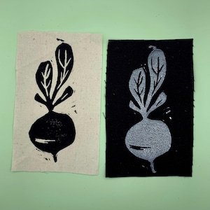 Op de afbeelding: Twee stoffen patches met een zwart-witte print van een raap. De raap is op een witte achtergrond op de linker patch en op een zwarte achtergrond op de rechter patch.