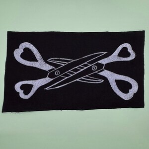 May include: Black rectangular patch featuring a white illustration of crossed scissors. The scissors have heart-shaped handles. The design is centered on the patch, with the scissors overlapping each other.